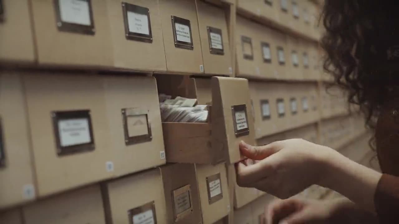 Two hands drawing open a wooden card-index drawer in a long wall of record drawers.